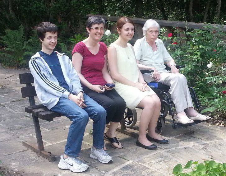 A young man and two ladies are sitting on a wooden bench. Another lady is sitting in a wheelchair next to one of the other ladies