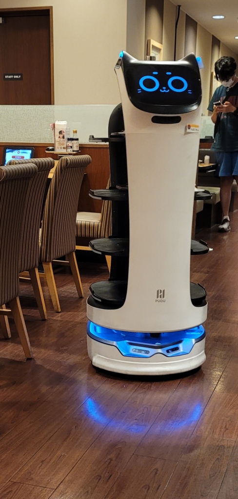 A white robot with four shelves delivering food. The screen on the robot was designed to look like a cartoon cat's face.